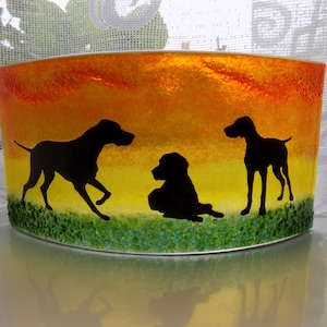 May include: A glass candle holder featuring a sunset scene with three black silhouettes of dogs. The base of the holder is green, the middle is yellow, and the top is orange.