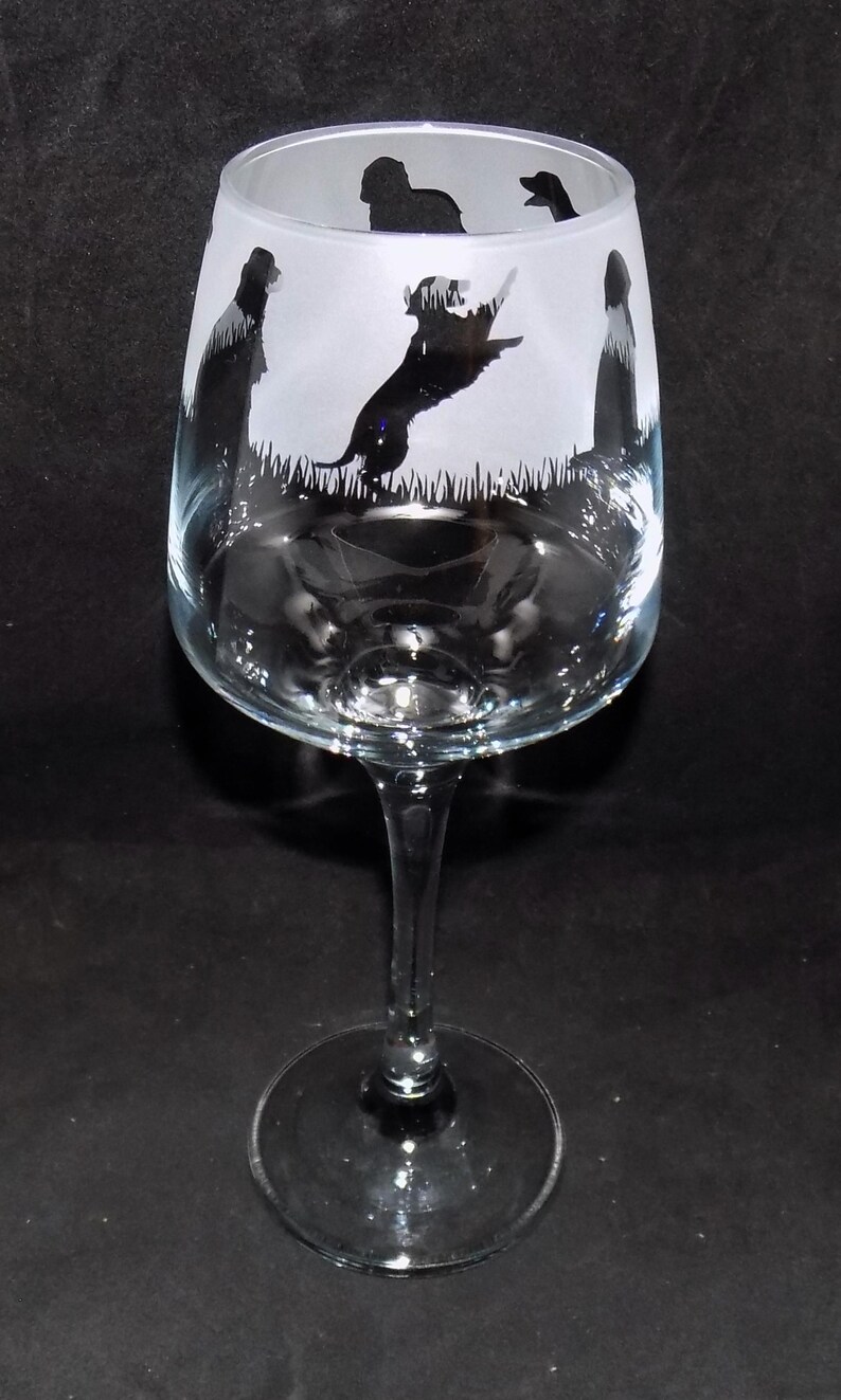 May include: A clear glass wine glass with a frosted design featuring silhouettes of dogs playing fetch. The glass has a long stem and a wide base.