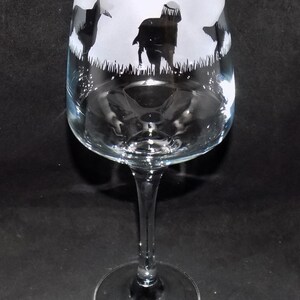 May include: A clear glass wine glass with a frosted design of four black silhouettes of dogs in a field of grass.