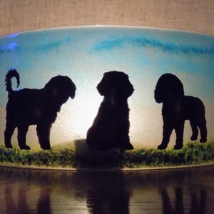 May include: A glass candle holder with a blue and white background featuring three black silhouettes of dogs. The dogs are standing on a green grass silhouette.