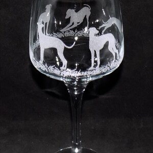 K&ouml;nnte beinhalten: Ein Weinglas aus klarem Glas mit ge&auml;tzten wei&szlig;en Silhouetten von mehreren Windhunden, die auf einer Wiese stehen und spielen. Der Stiel und der Fu&szlig; sind glatt und transparent und bilden einen Kontrast zu den detaillierten Gravuren.