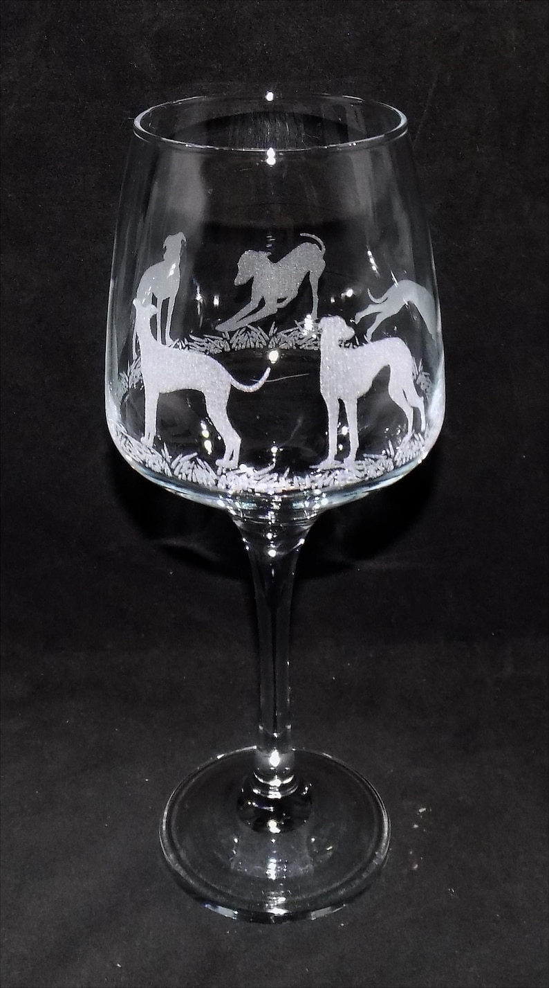 K&ouml;nnte beinhalten: Ein Weinglas aus klarem Glas mit ge&auml;tzten wei&szlig;en Silhouetten von mehreren Windhunden, die auf einer Wiese stehen und spielen. Der Stiel und der Fu&szlig; sind glatt und transparent und bilden einen Kontrast zu den detaillierten Gravuren.