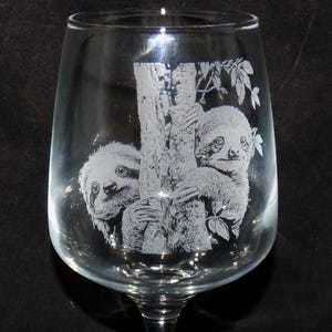 May include: Clear wine glass featuring an etched design of two sloths on a tree trunk. The sloths are intricately detailed. The glass has a long stem and a wide bowl, typical of a wine glass.