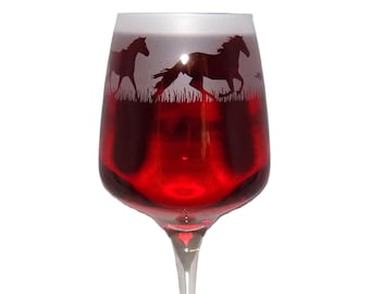May include: A clear glass wine goblet filled with deep red liquid, featuring a frosted upper band decorated with a dark silhouette of two horses running through a field of grass. The stem is clear and tapers down to a base.