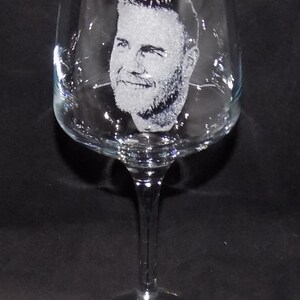 May include: A clear glass wine glass with a man's face etched into the side. The glass has a long stem and a round base.