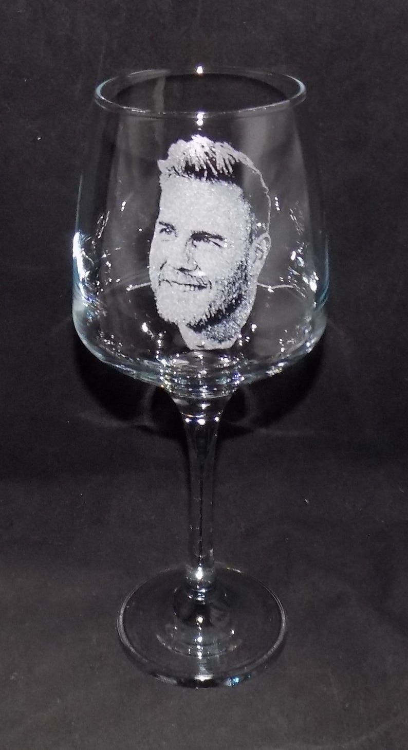 May include: A clear glass wine glass with a man's face etched into the side. The glass has a long stem and a round base.