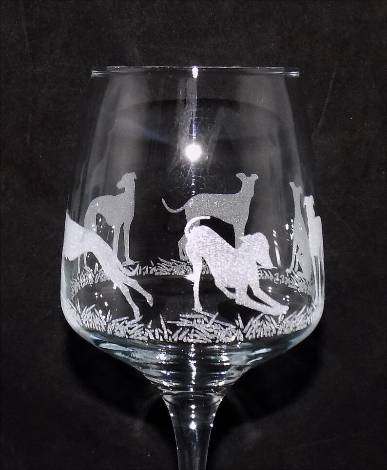 K&ouml;nnte beinhalten: Ein Weinglas aus klarem Glas mit einem ge&auml;tzten Design aus mehreren Windhunden in verschiedenen Posen, darunter stehend und sich streckend, vor einem texturierten Grasuntergrund. Die &Auml;tzung erscheint in einem mattwei&szlig;en Finish auf dem Glas.