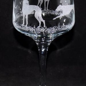 K&ouml;nnte beinhalten: Ein Weinglas aus klarem Glas mit einer ge&auml;tzten Darstellung mehrerer Windhunde in verschiedenen Posen, darunter stehend und rennend, vor einem texturierten Bodenmuster, das sich um die Kuppa des Glases auf einem schlanken Stiel erstreckt.