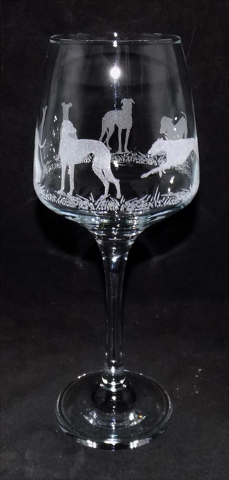K&ouml;nnte beinhalten: Ein Weinglas aus klarem Glas mit einer ge&auml;tzten Darstellung mehrerer Windhunde in verschiedenen Posen, darunter stehend und rennend, vor einem texturierten Bodenmuster, das sich um die Kuppa des Glases auf einem schlanken Stiel erstreckt.