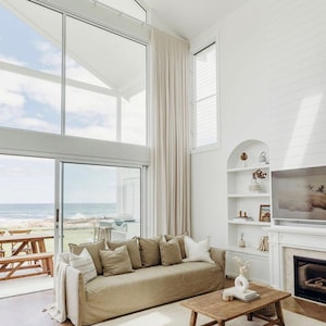 May include: A light brown linen sofa with multiple cushions sits in a living room with a large window overlooking the ocean. A wooden coffee table with a vase of flowers sits in front of the sofa. The room has white walls and a white rug.