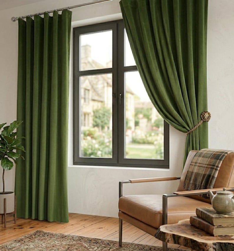 May include: Floor-length olive green velvet curtains hang from a silver rod over a window. One curtain panel is pulled back and secured with a gold-toned lion head tieback. The fabric features vertical pleats and a soft, matte texture.