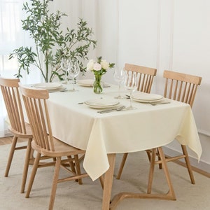Peut inclure: Une nappe blanche est posée sur une table en bois avec quatre chaises. La table est dressée avec des assiettes blanches, des couverts et des verres. Un vase avec des fleurs blanches est au centre de la table.