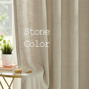 May include: A pair of sheer, stone-coloured curtains with a subtle textured weave. The curtains are hanging in a window and are partially drawn.