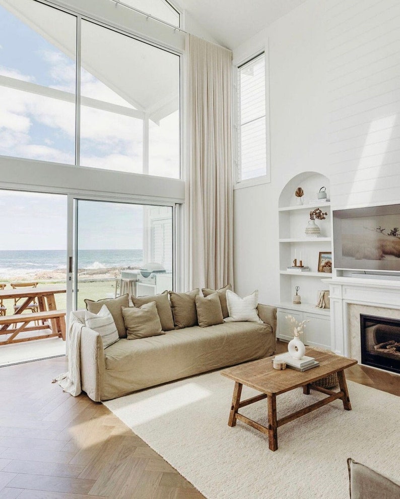 May include: A light brown linen sofa with multiple cushions sits in a living room with a large window overlooking the ocean. A wooden coffee table with a vase of flowers sits in front of the sofa. The room has white walls and a white rug.