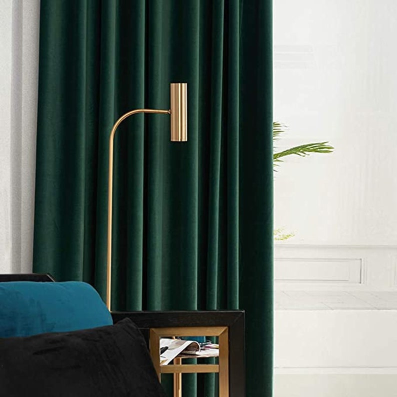 May include: A floor lamp with a thin gold-toned metal stand and a cylindrical head, positioned in front of deep green velvet curtains. A black and gold side table is partially visible, along with a teal and a black cushion in the foreground.