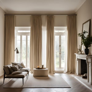 May include: Floor-to-ceiling beige curtains hang from a black rod over two white French doors. A beige sofa with matching throw pillows sits on a light rug next to a round beige ottoman. A fireplace with a mirror and a black vase is on the right.
