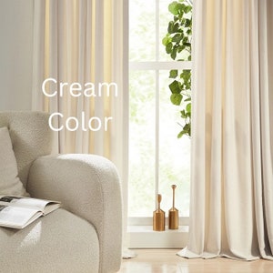 May include: Cream coloured curtains hanging in a window with a plant in front of the window. The curtains are made of a soft, flowing fabric. The text "Cream Color" is written on the curtains.