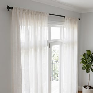 Pu&ograve; includere: Una coppia di tende semitrasparenti color bianco sporco appese a un bastone in metallo scuro con terminali decorativi. Le tende presentano una tasca per il bastone e cadono in morbide pieghe verticali, lasciando filtrare la luce attraverso.