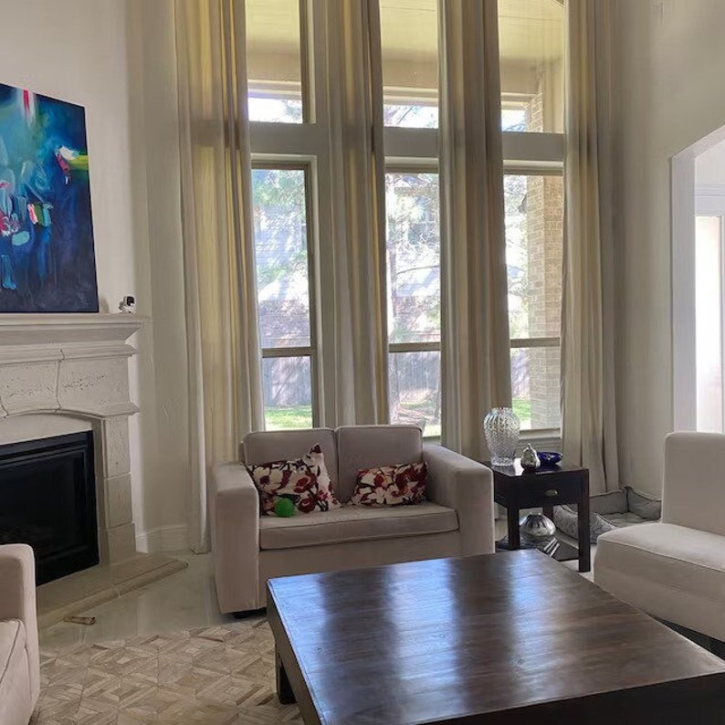 May include: A living room featuring a beige loveseat with two floral patterned throw pillows, a dark wood square coffee table, a stone fireplace with an abstract blue painting above it, and tall windows covered by floor-to-ceiling beige curtains.