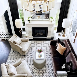 May include: A living room featuring a dark brown sofa with assorted throw pillows, two beige armchairs, and a gray oval ottoman. A large gold chandelier hangs above a fireplace with a mirror. A geometric patterned rug covers the dark wood floor.
