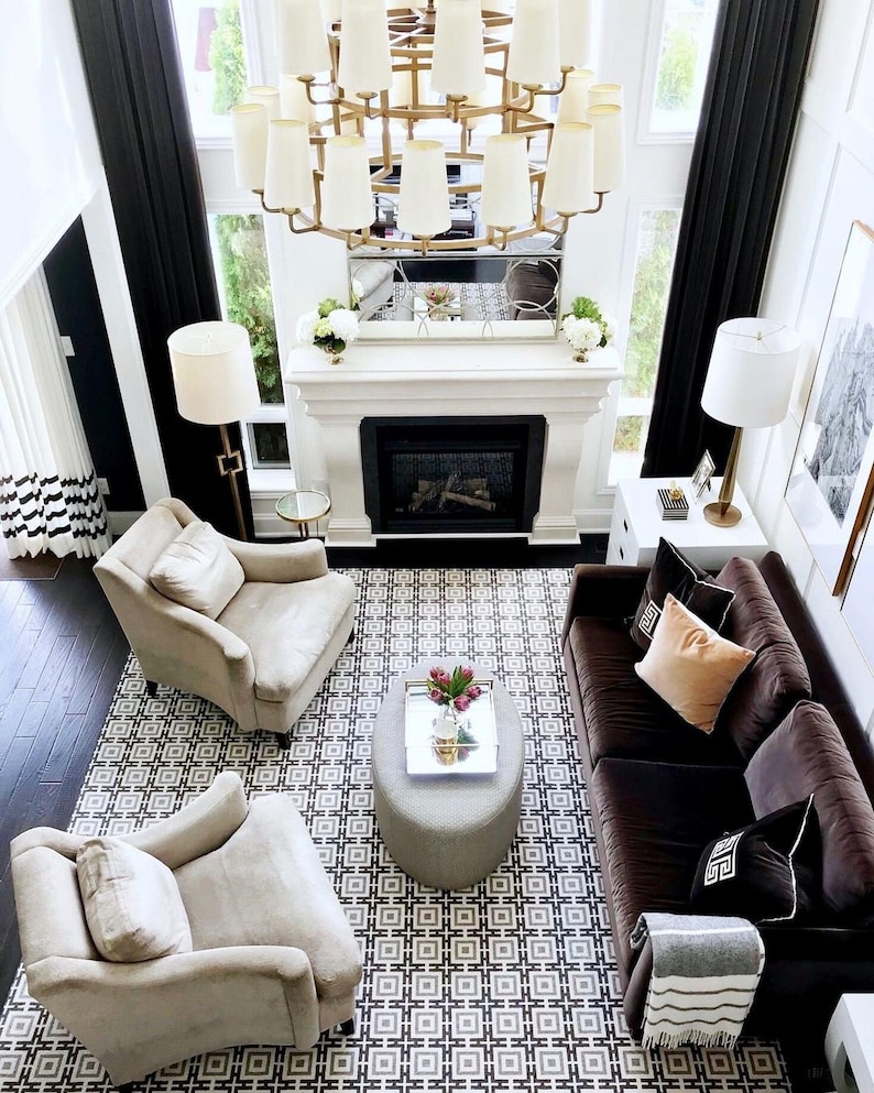 May include: A living room featuring a dark brown sofa with assorted throw pillows, two beige armchairs, and a gray oval ottoman. A large gold chandelier hangs above a fireplace with a mirror. A geometric patterned rug covers the dark wood floor.