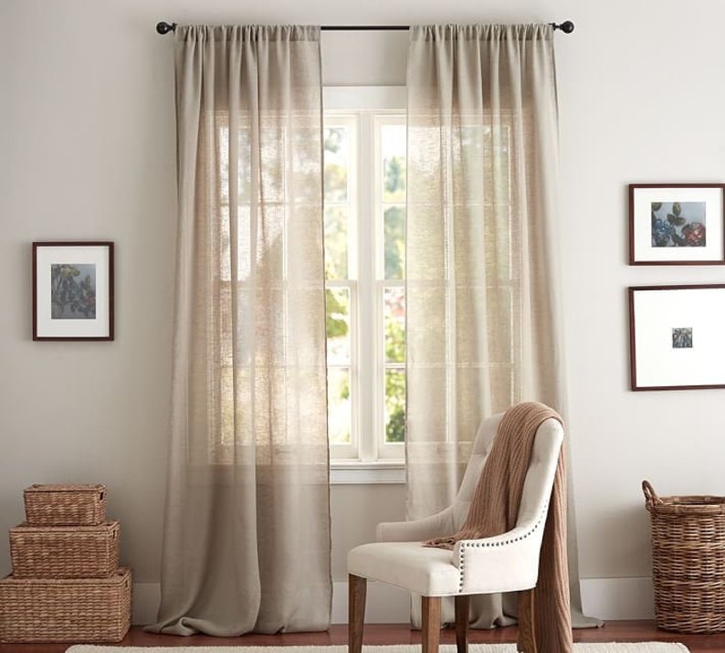 Pu&ograve; includere: Una coppia di tende beige semitrasparenti che arrivano fino a terra, appese a un bastone nero sopra una finestra. Le tende presentano una trama visibile e sono posizionate dietro una sedia imbottita color bianco sporco con un plaid.