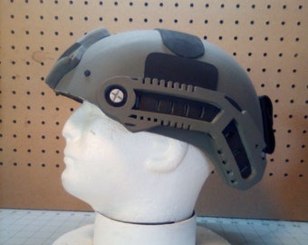 May include: A grey tactical helmet with a black padded headband and a black strap. The helmet has a black and grey rail system on the side.