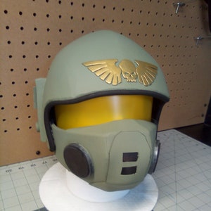 Puede incluir: Un casco verde y dorado con un emblema de calavera y alas. El casco tiene una visera amarilla y orejeras negras.