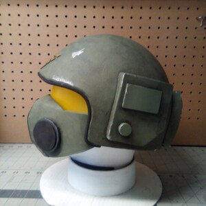 May include: A green and gray helmet with a yellow visor and a black face mask. The helmet has a rectangular panel on the side with a small round button.