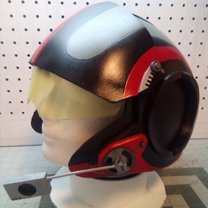 May include: A black, red, and gray helmet with a clear visor and a microphone. The helmet is on a white stand.