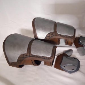 May include: Two brown leather and silver metal arm guards with two additional silver metal pieces. The arm guards have a textured design.