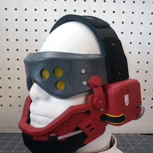 May include: A red and gray 3D printed helmet with a black strap and yellow accents. The helmet has a visor with multiple holes and a red earpiece with a lever.