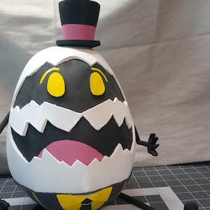 Może przedstawiać: Biała postać w kształcie jajka z czarnym kapeluszem, czarnym krawatem i żółtym krawatem. Postać ma czarno-białą twarz z żółtymi oczami i różowymi ustami. Postać ma dwie czarne ręce i nogi.