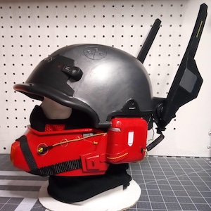 May include: A black and red helmet with a gray helmet shell, a red face mask, and a black antenna on the back. The helmet has a black strap and a black and red design.