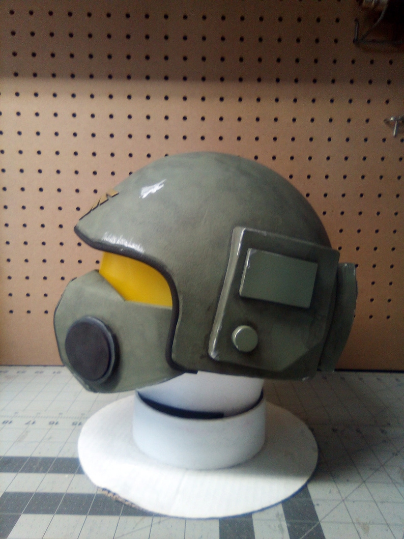 May include: A green and gray helmet with a yellow visor and a black face mask. The helmet has a rectangular panel on the side with a small round button.