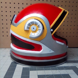 May include: A red, yellow, silver, and black helmet with a large circular detail on the side. The helmet has a visor and is made of a hard plastic material.
