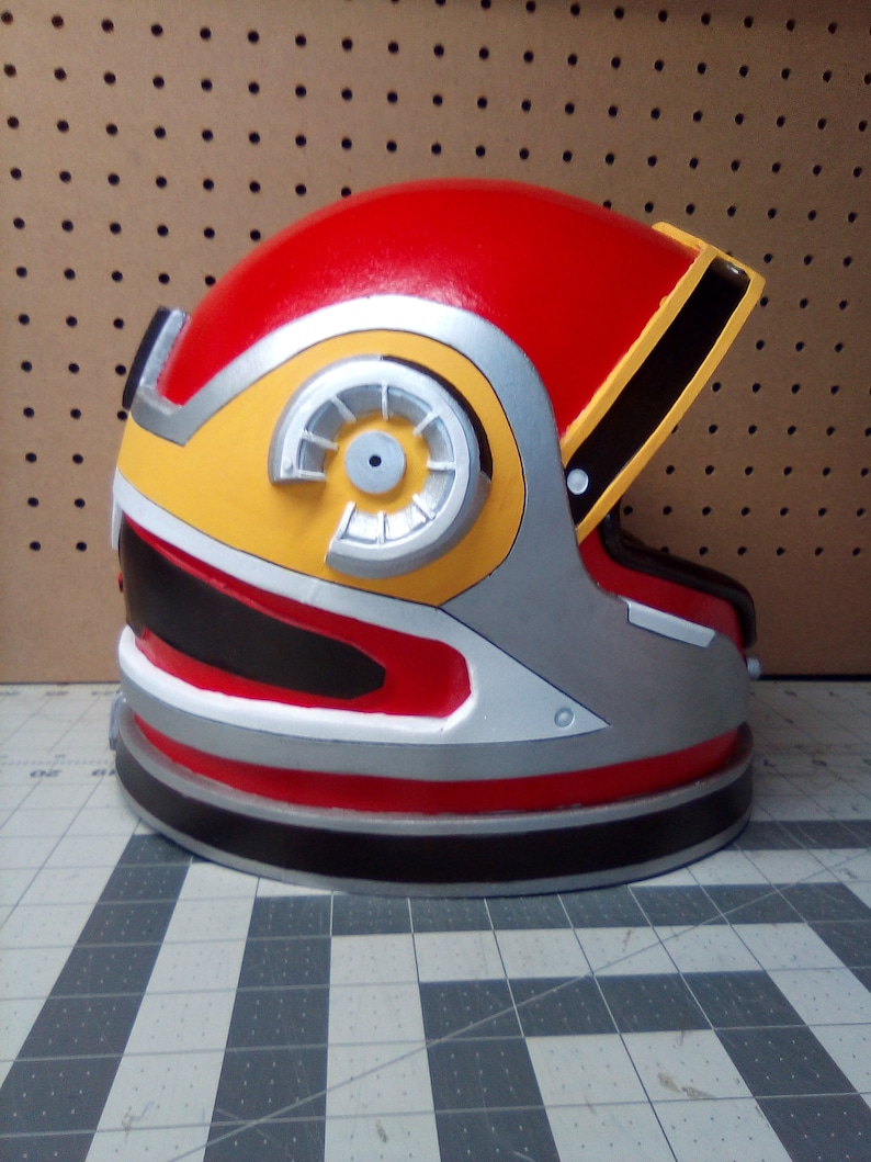 May include: A red, yellow, silver, and black helmet with a large circular detail on the side. The helmet has a visor and is made of a hard plastic material.