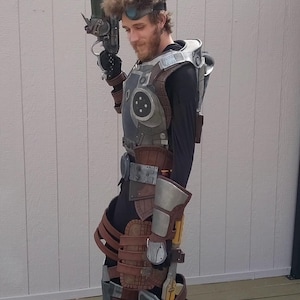 Peut inclure: Un costume de cosplay détaillé avec un design futuriste. La tenue comprend une pièce de poitrine métallique, des accents marron et gris, et de grandes bottes. La personne tient une arme factice, ce qui ajoute au thème de la science-fiction.