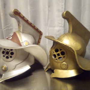 Puede incluir: Dos cascos de gladiador, uno plateado y otro dorado, con detalles intrincados y una cresta elevada en la parte superior.