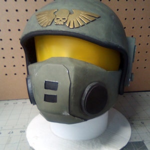 May include: A green and gold helmet with a skull and wings design. The helmet has a yellow visor and a black face mask.