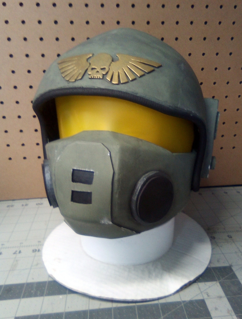 May include: A green and gold helmet with a skull and wings design. The helmet has a yellow visor and a black face mask.