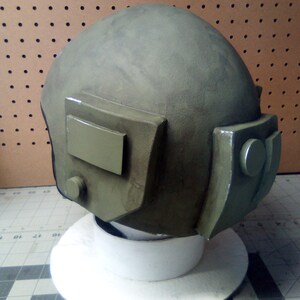May include: A green military helmet with a visor and a small round button on the side. The helmet is sitting on a white base.