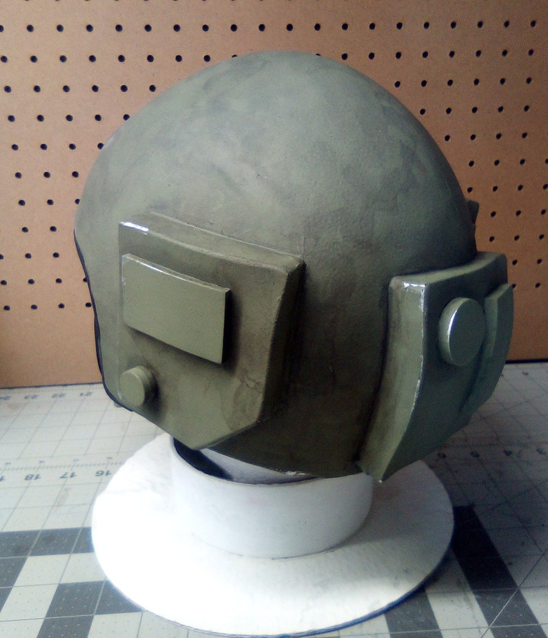 May include: A green military helmet with a visor and a small round button on the side. The helmet is sitting on a white base.