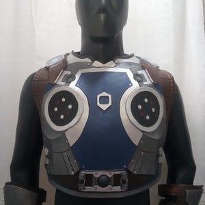 May include: A futuristic chest armour and arm guards in shades of blue, silver, and brown, displayed on a mannequin. The armour features circular accents, a central blue panel, and is paired with blue-tinted goggles.
