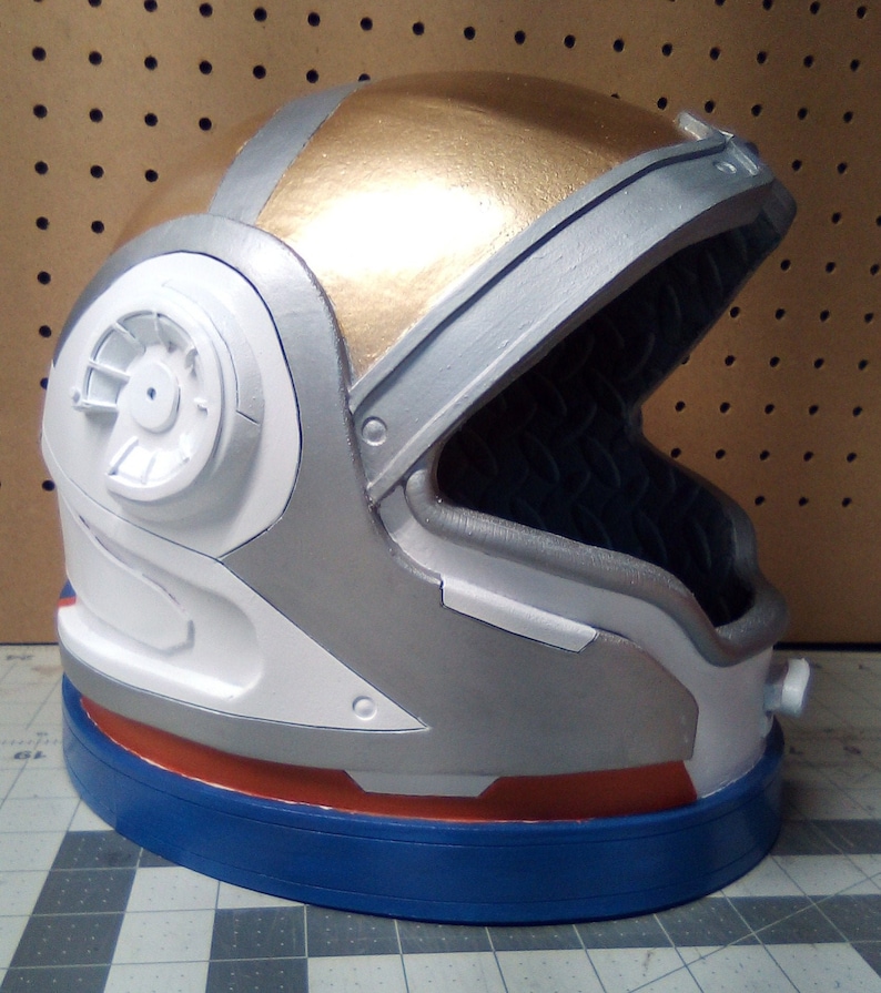 May include: A white, silver, and gold astronaut helmet with a blue and orange trim. The helmet has a visor that is closed and a small round window on the side.