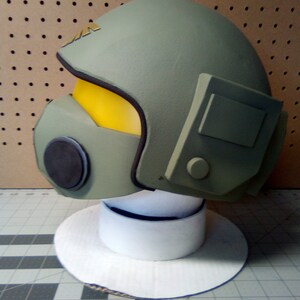 May include: A green helmet with a yellow visor and a black microphone. The helmet has a white base and is on a white circular stand.