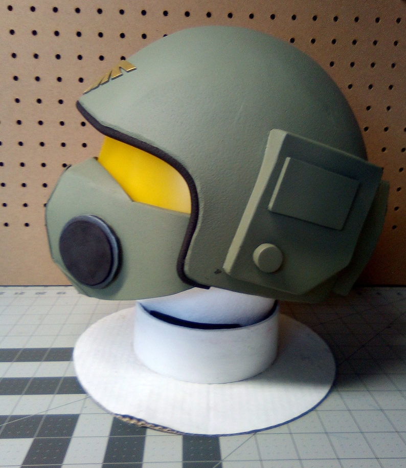 May include: A green helmet with a yellow visor and a black microphone. The helmet has a white base and is on a white circular stand.