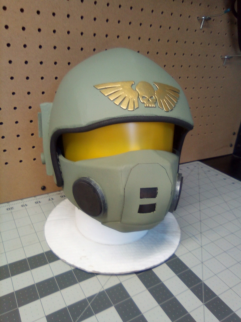 May include: A green and gold helmet with a skull and wings emblem. The helmet has a yellow visor and black earpieces.