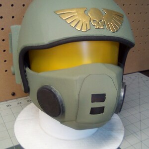May include: A green and brown helmet with a gold skull and wings emblem. The helmet has a yellow visor and a black microphone.