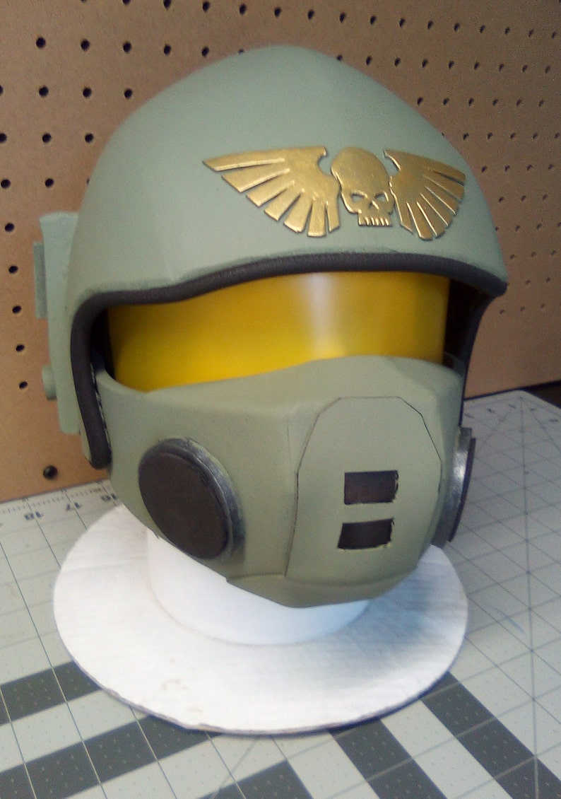 May include: A green and brown helmet with a gold skull and wings emblem. The helmet has a yellow visor and a black microphone.